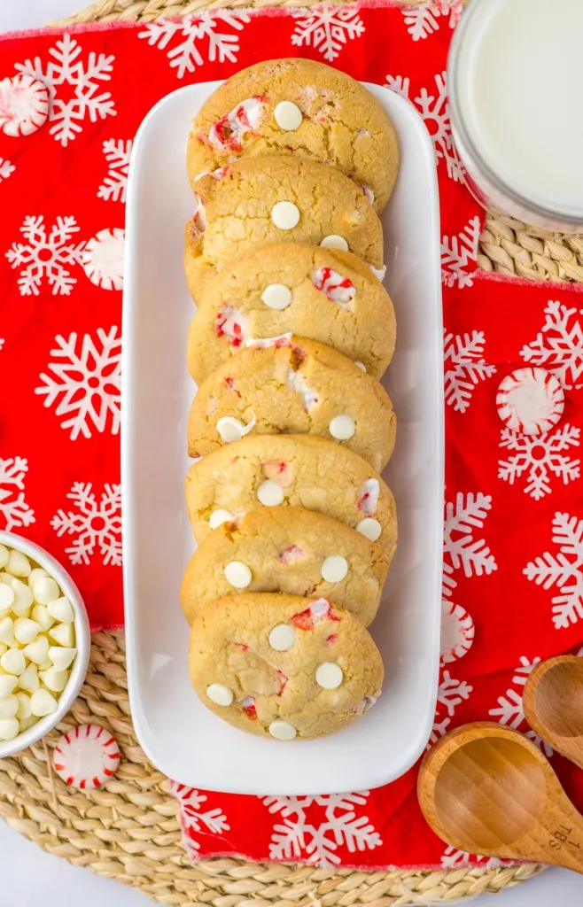 Overhead shot of baked white chocolate peppermint cookies on a white tray, ingredients surrounding, on a holiday tablecloth.