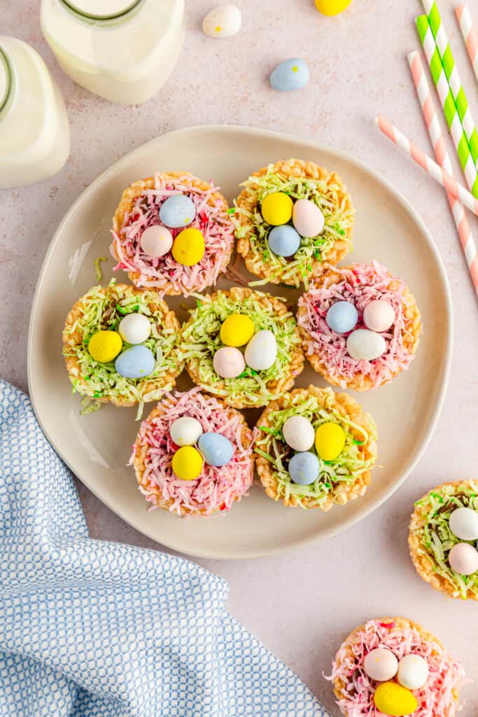 Overhead view of Rice Krispies Easter Nests on a serving plate with jars of milk, pastel blue kitchen cloth, mini eggs candy, and striped straws on a countertop. 