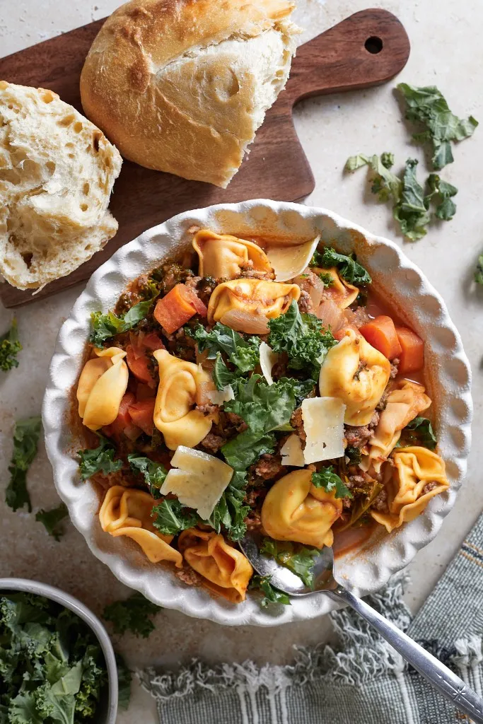Sausage Kale Tortellini Soup in a white soup bowl, wooden kitchen board with bread, small bowl of kale.