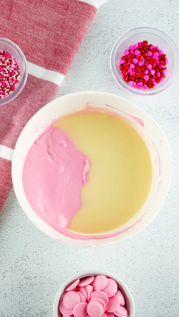 Overhead view of a bowl with pink melted candy melts and heated buttercream mixture, small bowl of pink candy melts, and sprinkles.