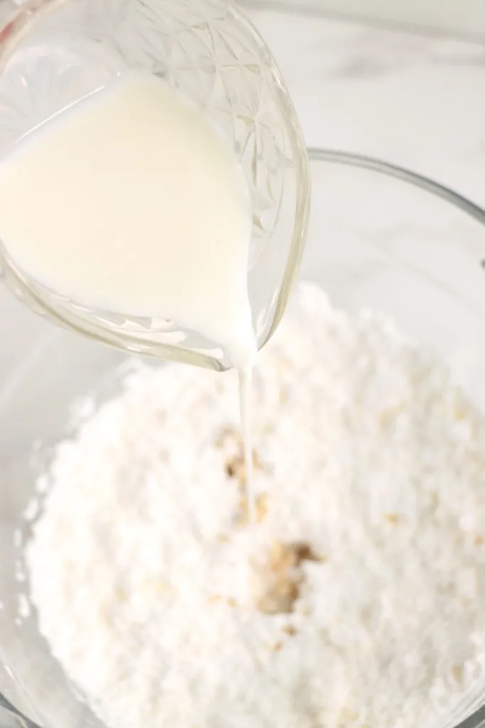 Glass bowl with vanilla buttercream frosting ingredients and milk being poured into the mixture.