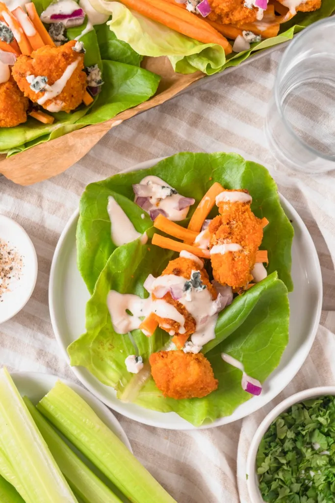 Overhead shot of assembled Buffalo Chicken lettuce wrap on a white serving plate, glass of water, and extra lettuce wraps in the background.