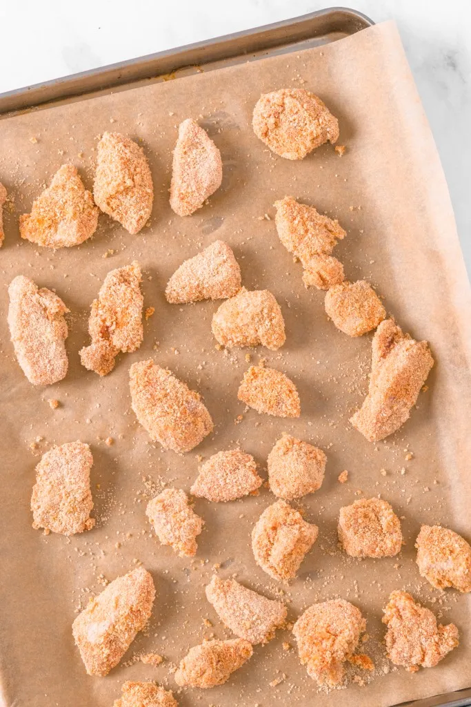 Baking tray lined with parchment paper and filled with marinated chicken pieces that are coated in breadcrumbs. 