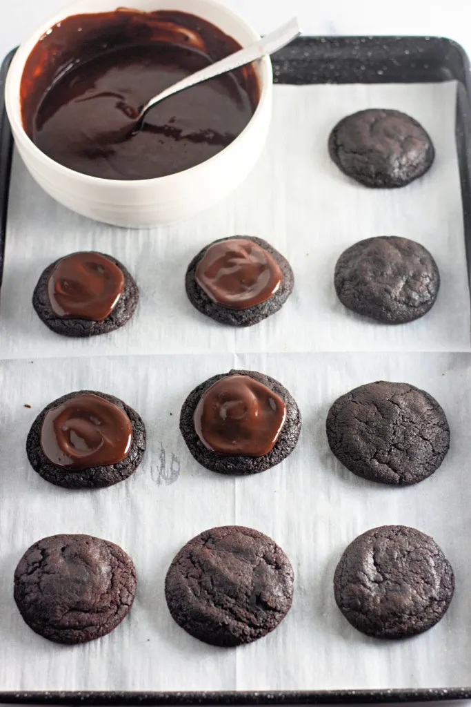 Baking tray with baked cosmic brownie cookies, chocolate frosting being spread over the top of each cookie in preparation of adding sprinkles.