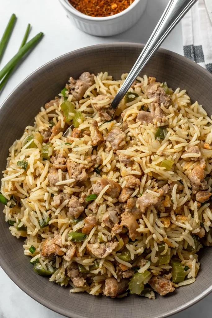 Close up of Dirty Rice with Sausage in a serving bowl with a spoon, ingredients in the background with a striped kitchen cloth. 