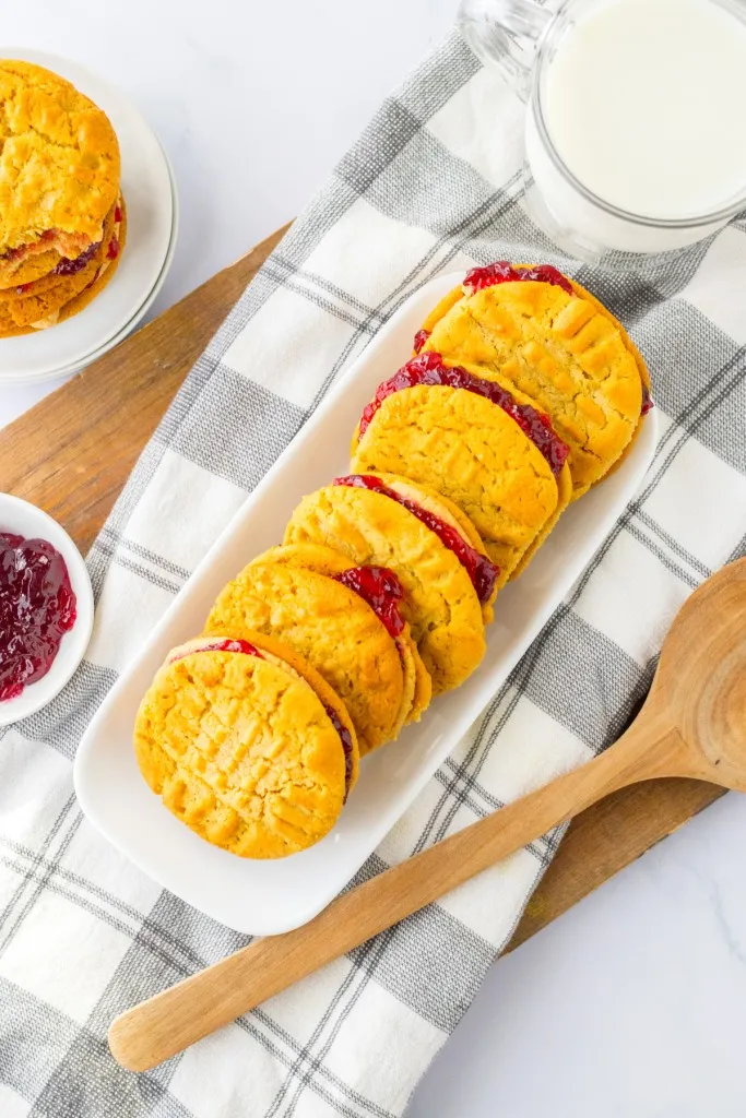 Tray of peanut butter and jelly cookie sandwiches, stacked cookies on a white plate, small bowl of grape jelly, wooden spoon on a kitchen cloth.