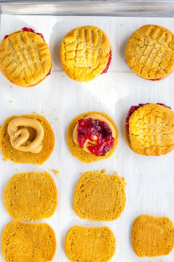Peanut cookie halves with piped buttercream and grape jelly on a baking sheet to assemble peanut butter and jelly cookie sandwich.
