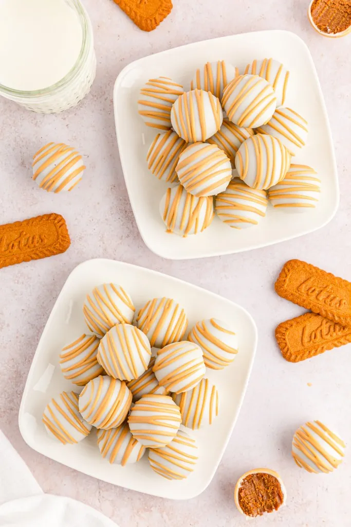 Biscoff truffles piled on two white plates. Biscoff cookies and truffles lying on a countertop, one Biscoff truffle bitten in half.