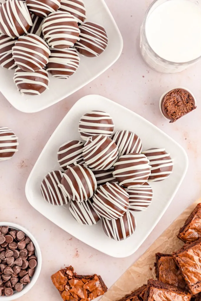 Overhead shot of assembled brownie balls on two white serving plates, small bowl of dark chocolate melting wafers, glass of milk, and brownie squares.