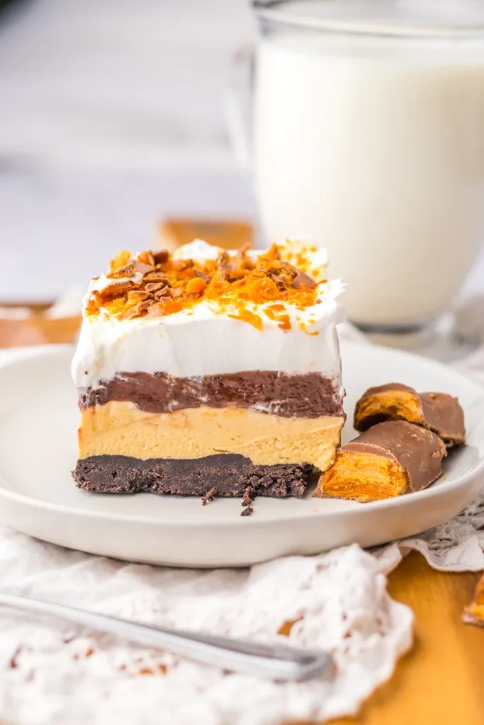 Close up of a slice of Butterfinger lush dessert on a white plate with additional Butterfinger candy.