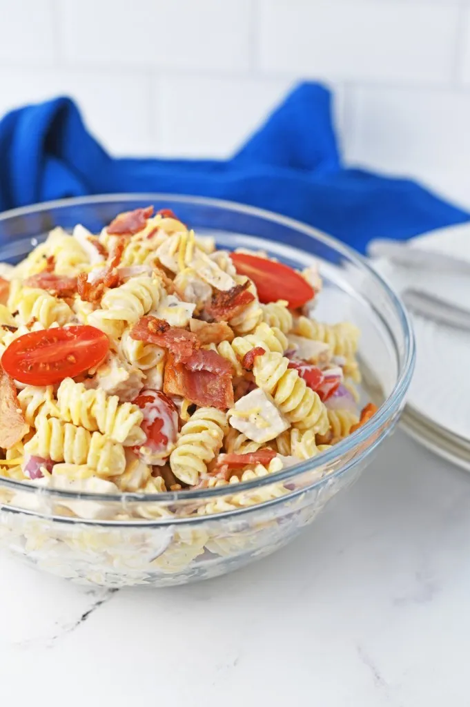 Close up of chicken bacon ranch pasta salad on a marble countertop, blue kitchen cloth, white serving plates, and metal utensils in the background.