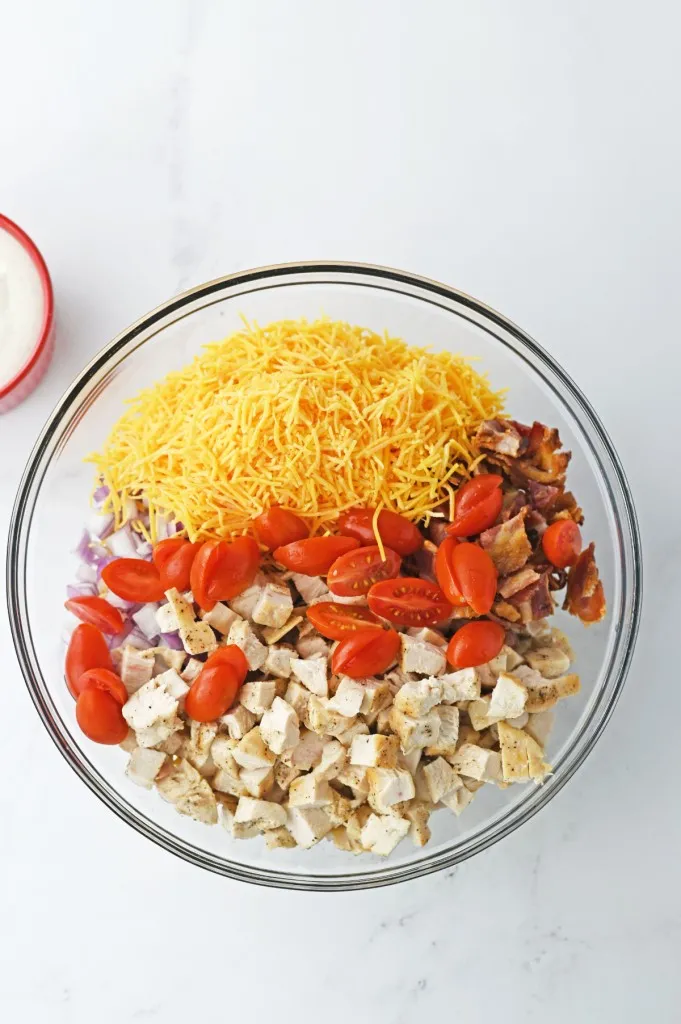 A large glass mixing bowl with freshly shredded cheese, halved cherry tomatoes, chopped chicken, chopped red onion, and roughly chopped bacon.