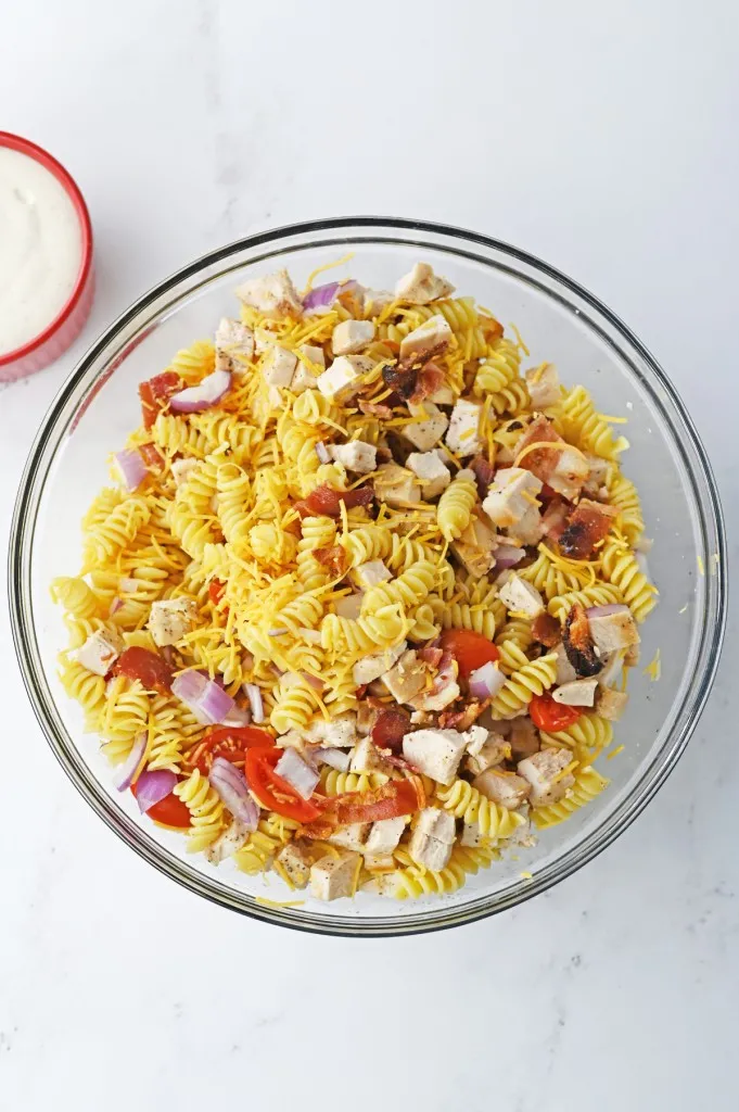 Large glass mixing bowl with chopped chicken, bacon, red onions, halved tomatoes, shredded cheese, and rotini pasta, all tossed together.