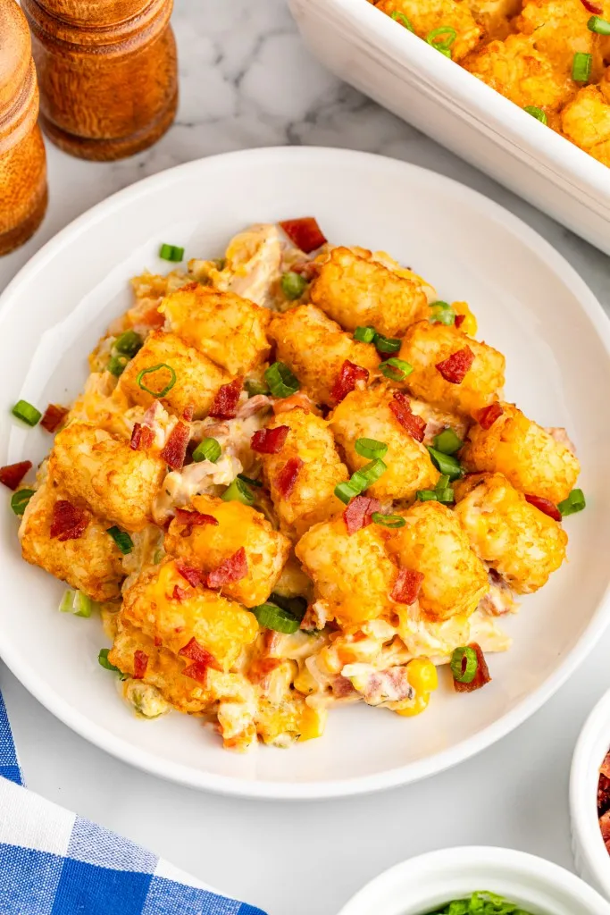 Serving of chicken tater tot casserole on a white plate, rest of the casserole in a white casserole dish, wooden salt and pepper shakers, blue and white checkered cloth, and silver fork.