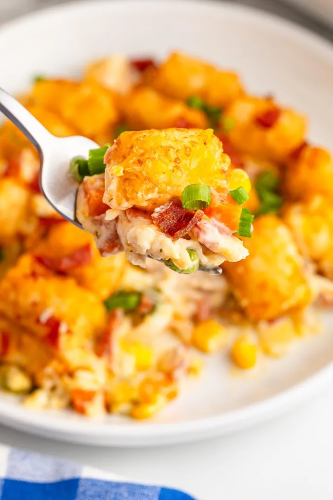 Fork loaded with chicken tater tot casserole above a serving of casserole on a white plate.