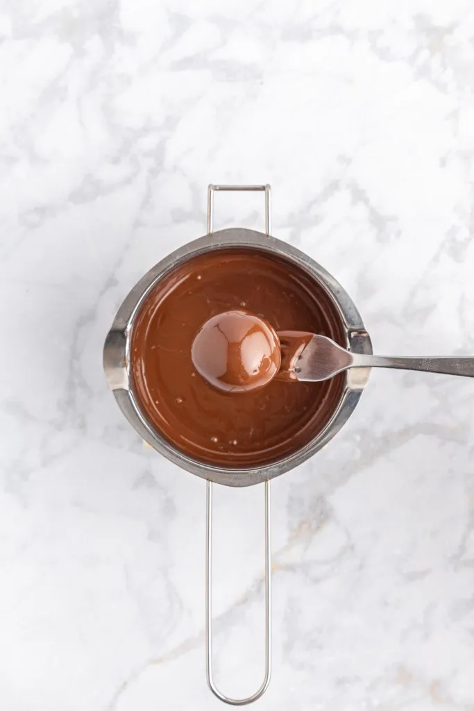 Double boiler with melted dark melting wafers, peanut butter ball sitting on a metal fork and fully coated in dark chocolate.