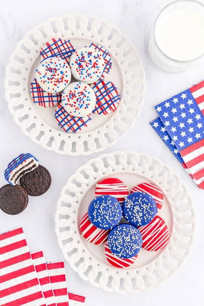 Overhead view patriotic Oreo cookies on white decorative plates, one Oreo cookie bitten into, patriotic-themed napkins on a white countertop.