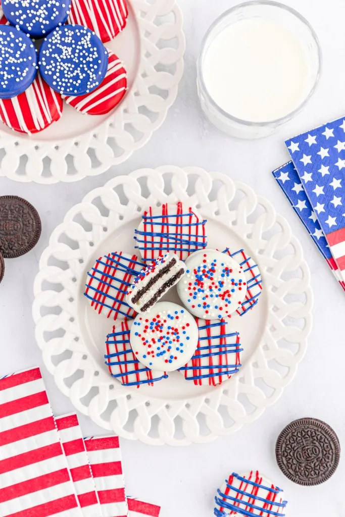 Overhead view of decorated patriotic Oreo cookies, piled on white serving plates, glass of milk, patriotic-themed napkins on a white countertop.