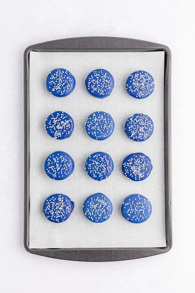 Overview of a baking tray lined with parchment paper and Oreo cookies that have been coated in melted blue candy melts with white nonpareils.