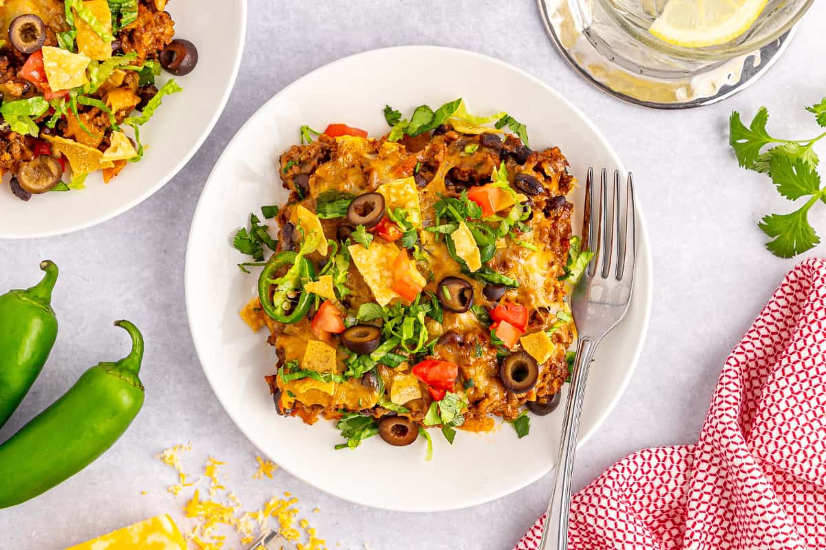 Overhead view of a white dinner plate with a serving of easy taco casserole, glass of water, red checkered kitchen cloth, and casserole ingredients in the background.