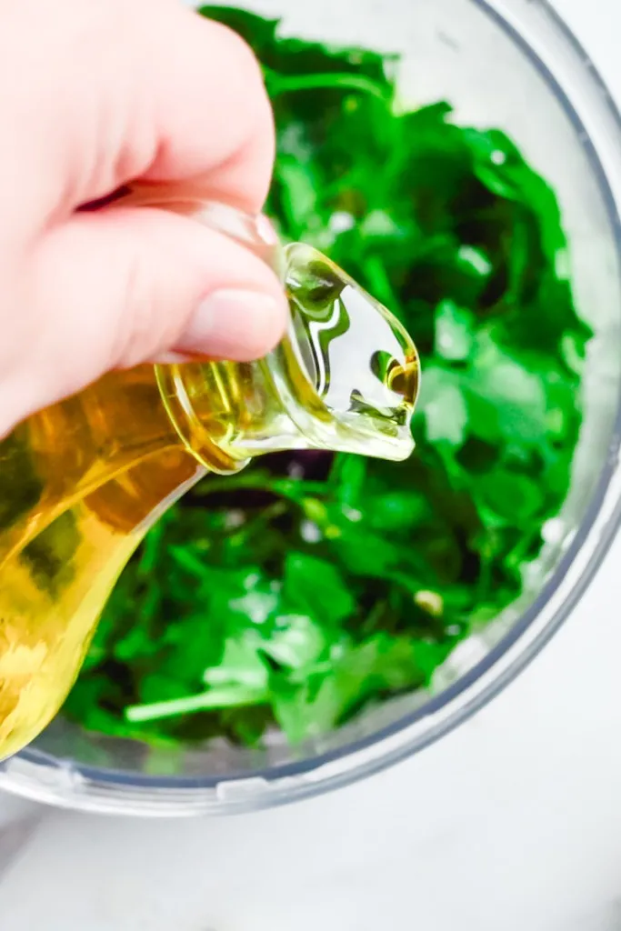 Hand pouring olive oil into a food processor with parsley, garlic, lemon juice, and lemon zest.