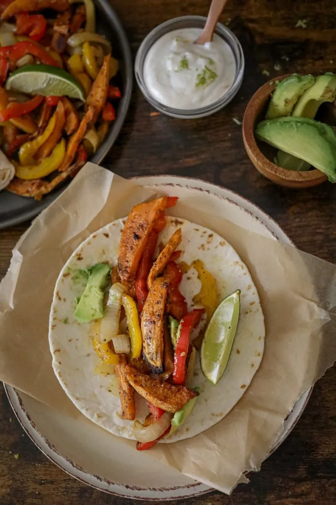 Vegetables with a wedge of lime on top of a heated tortilla on a serving plate, sliced avocado, zesty lime sauce, and roasted veggies in the background.