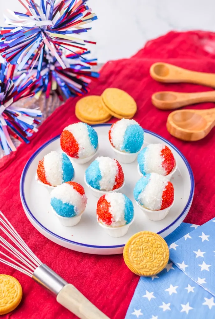 Snow cone Oreo truffles on a white plate on a red tablecloth, decorative napkins, metal whisk, wooden spoons, and loose Golden Oreos.