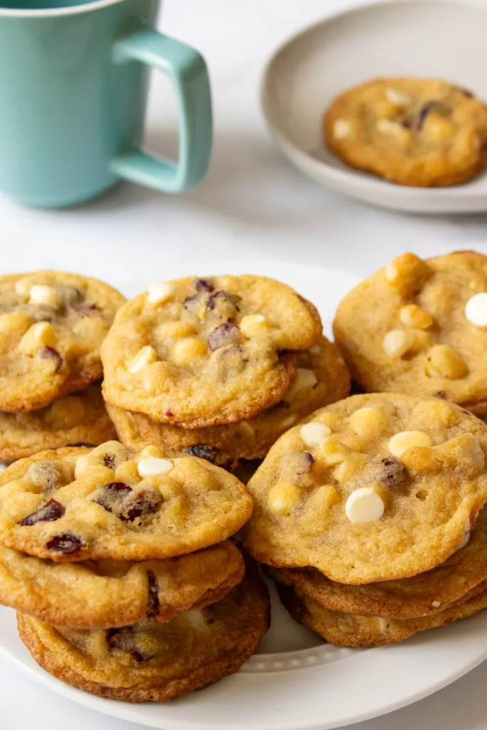 White chocolate cranberry hazelnut cookies stacked on a white plate, a hot beverage in a mug in the background.