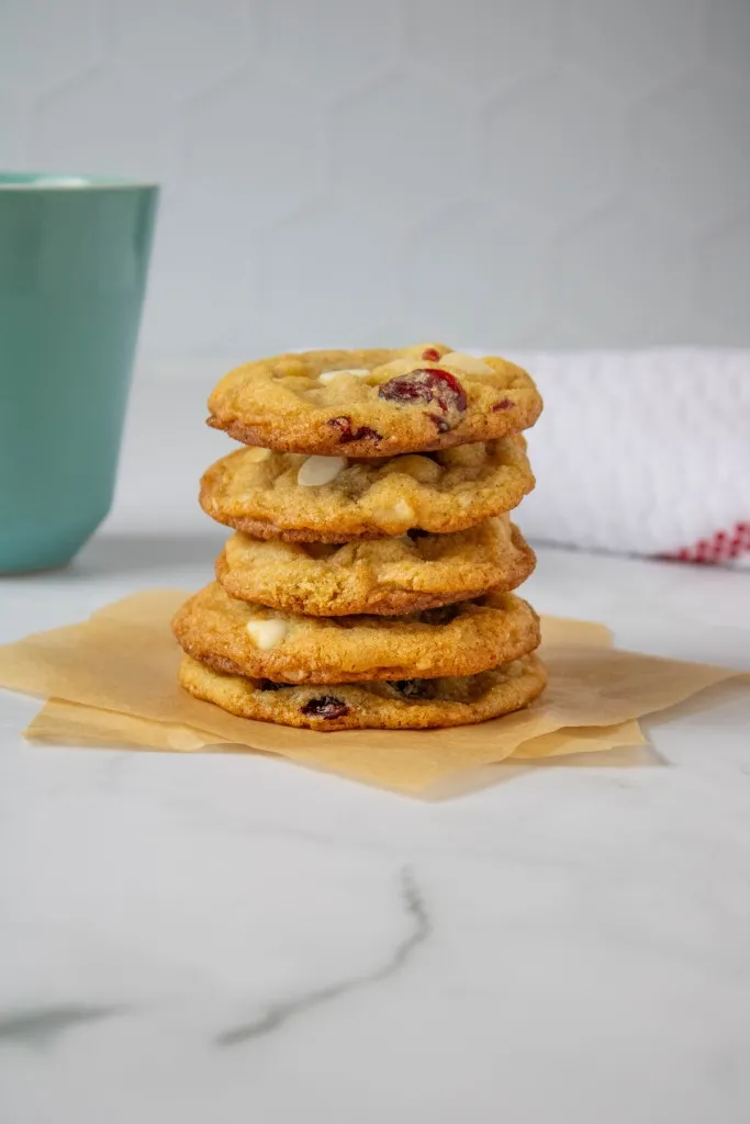 Stacked white chocolate cranberry hazelnut cookies on squares of parchment paper, hot beverage in the background with a white kitchen cloth in the background.