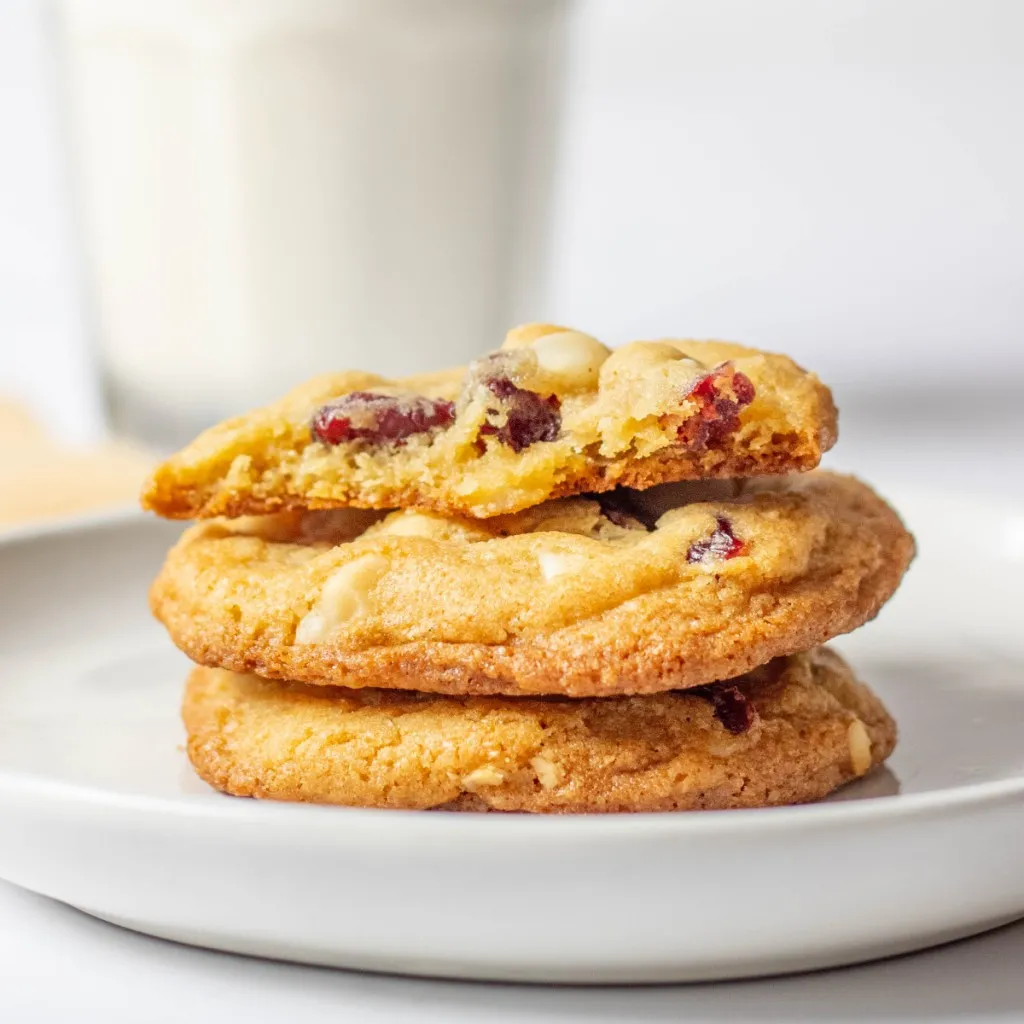 Stacked white chocolate cranberry hazelnut cookies on a white plate with a glass of milk behind.