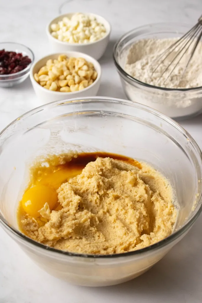 Glass mixing bowl with creamed butter, sugar, vanilla, egg, and yolk. Separate mixing bowl in the background with dry ingredients, white ramekins with white chocolate chips, nuts, and dried cranberries.
