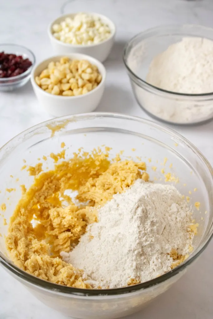 Glass mixing bowl with wet cookie dough mixture and dry ingredients. Separate mixing bowl with dry ingredients, white ramekins with white chocolate chips, nuts, and dried cranberries.