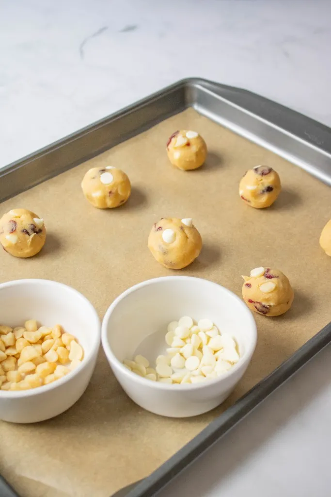 Cookie dough balls on a parchment-lined baking sheet, white ramekins with white chocolate chips and nuts.