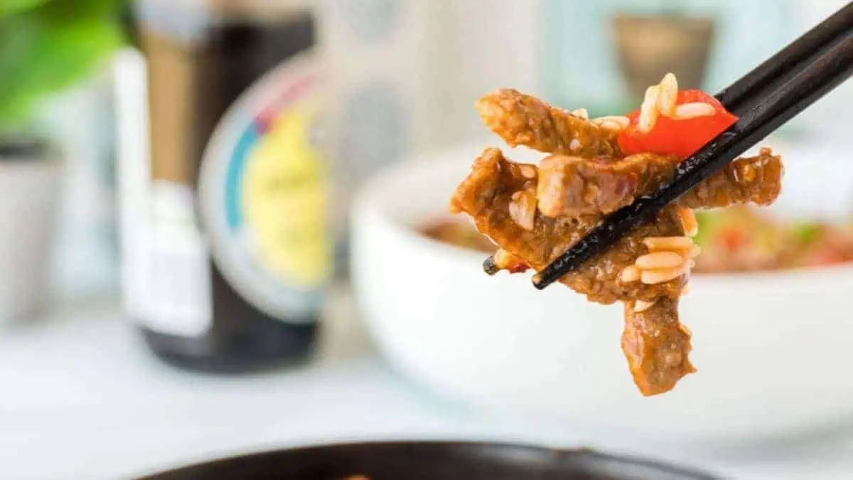 Chopsticks lifting tender, saucy strips of beef and peppers from a plate.