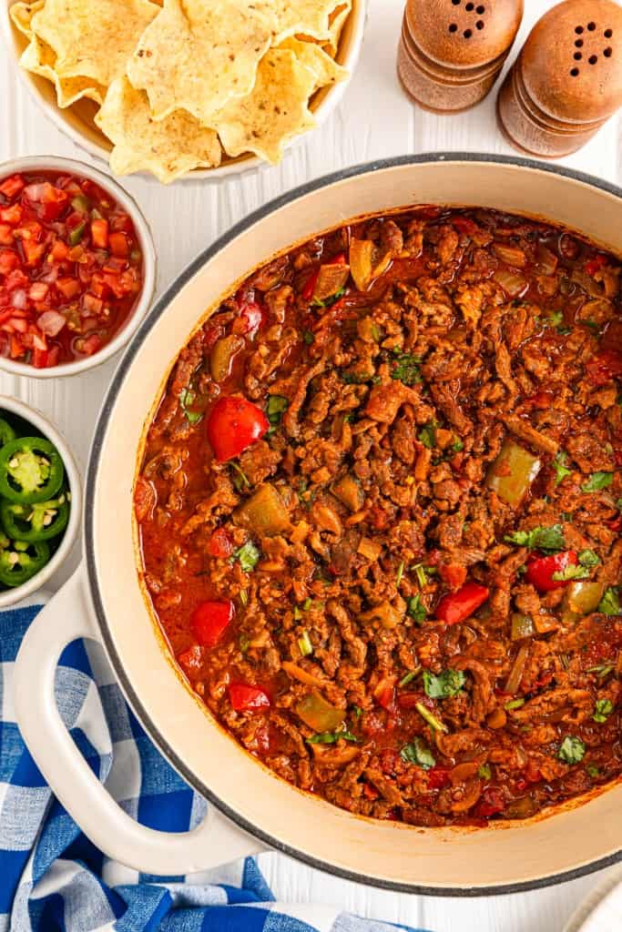 Carne Picada in a Dutch Oven. Canned diced tomatoes, chopped green bell peppers, and tortilla chips in separate bowls. Salt and pepper shakers to the side of the ingredients. 