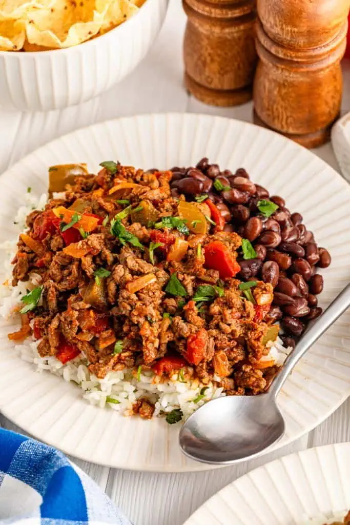 A plate of carne picada served over rice with a metal spoon, salt and pepper shakers in the background with a bowl of tortilla chips. 