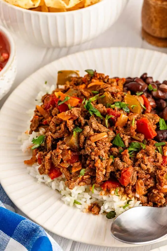 Close up of a serving of Carne Picada served over white rice, sprinkled with fresh chopped cilantro on a white plate with a metal spoon. 