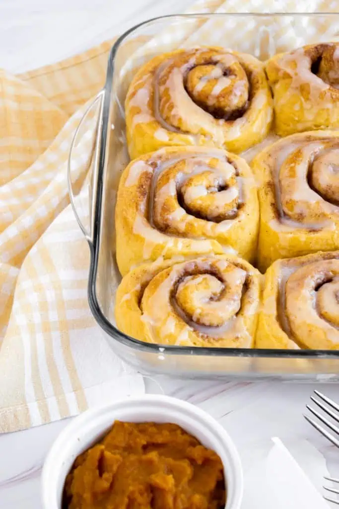 Pumpkin cinnamon rolls in a glass baking dish, drizzled with a sugar glaze, white ramekin with pumpkin puree, orange checkered kitchen cloth, and two sliver forks to the side.