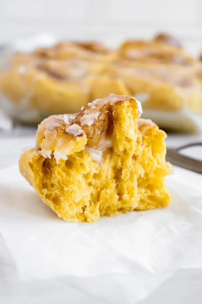 Close up of a pumpkin cinnamon roll that has been pulled in half, the remainder of the baked pumpkin cinnamon rolls in a glass baking dish in the background.