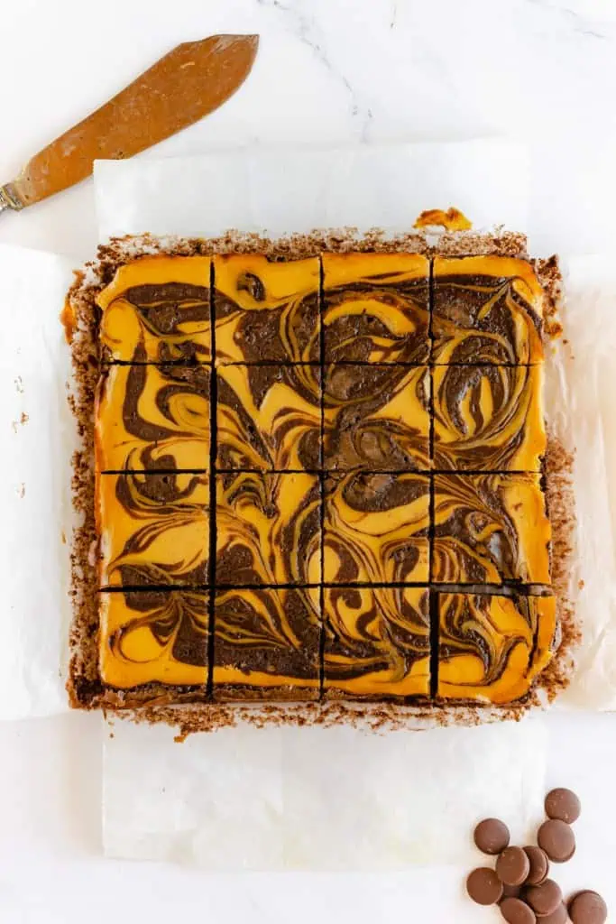 Sliced pumpkin cream cheese brownies on parchment paper with metal serving knife and scattered chocolate chips.