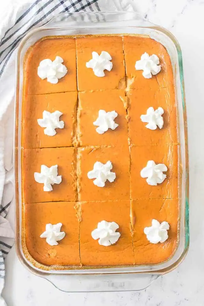 Overhead view of pumpkin pie dessert bars in a glass baking dish, topped with small dollops of whipped cream, blue and white striped cloth on a marble countertop.