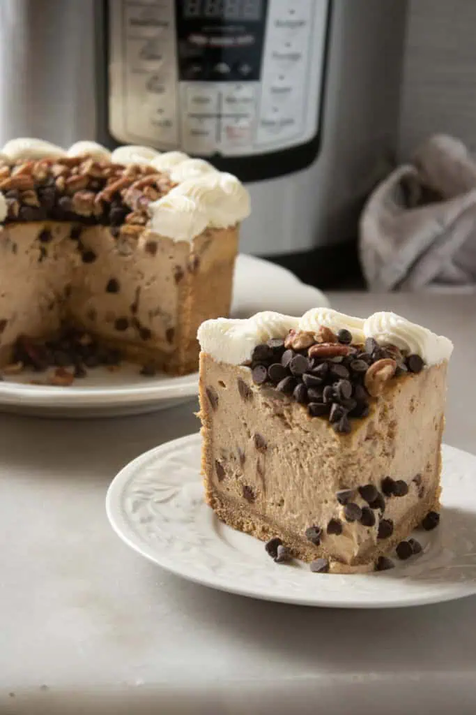 A slice of Instant Pot Pumpkin Chocolate Chip Cheesecake on a white serving plate, the remainder of the cheesecake on a large serving plate in the background.