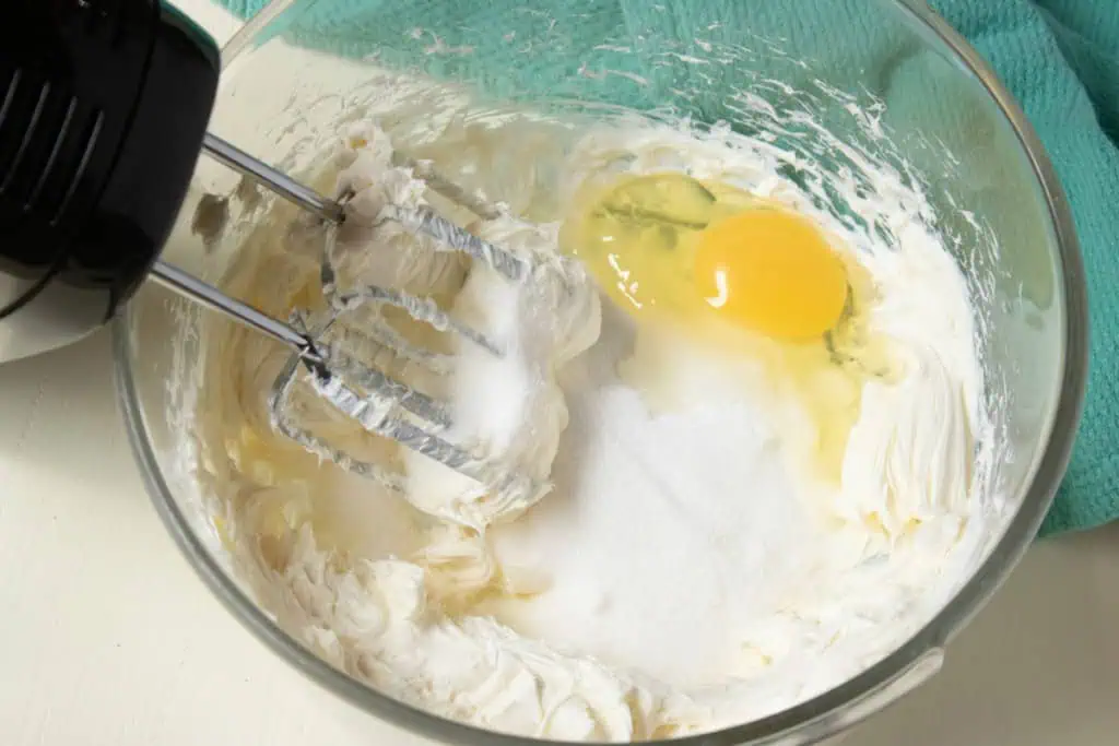 Glass mixing bowl with softened cream cheese, granulated sugar, and eggs. Electric mixer with attachments covered with cream cheese mixture.