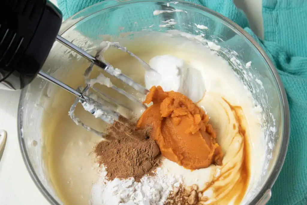 Glass mixing bowl with cream cheese mixture, sour cream, flour, pumpkin puree, pumpkin spice, and vanilla extract. Electric mixer with attachments in the bowl.