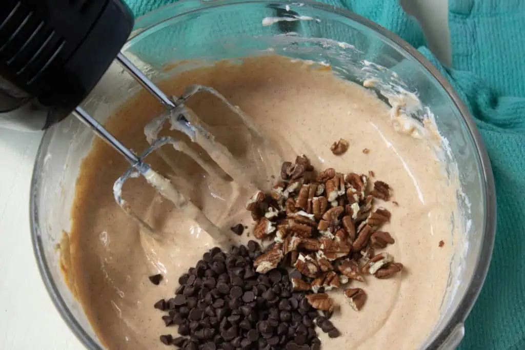 Glass bowl with cheesecake filling, half of the chocolate chips and chopped pecans added, electric mixer with attachments in the bowl.