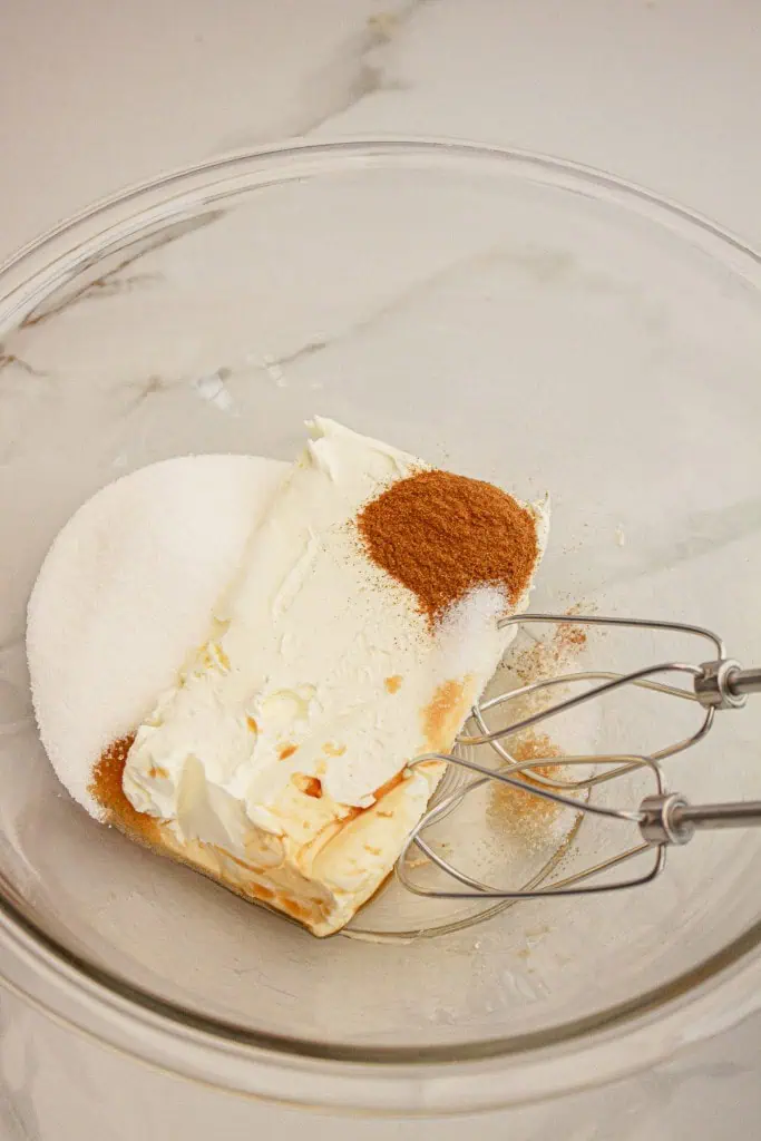 A large mixing bowl with cream cheese, sugar, vanilla, salt, and pumpkin spice with electric beater metal attachments. 