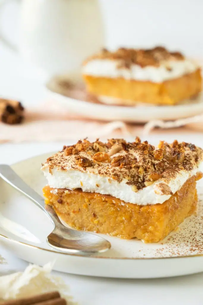 Close up of a slice of sweet potato pudding on a white serving plate with metal spoon. Another slice of sweet potato pudding in the background.
