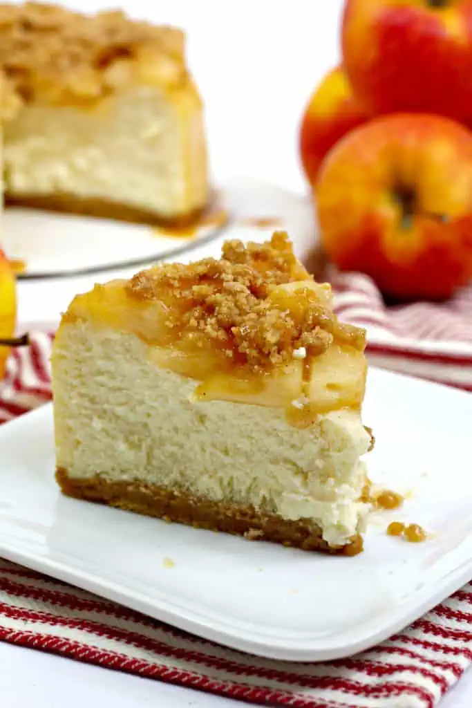Close up of a slice of Instant Pot apple crumble cheesecake on a white serving plate, whole cheesecake with apples in the background.