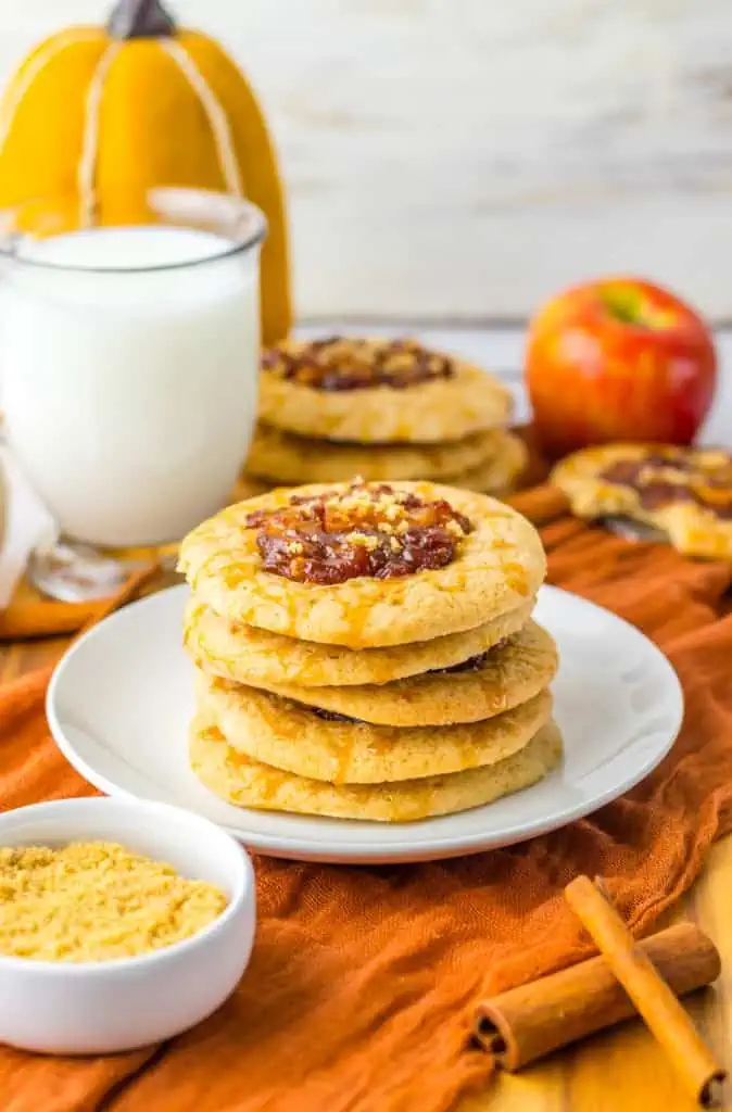 Close up apple spiced cookies stacked on a white plate, glass of milk, whole apple, and additional cookies in the background.