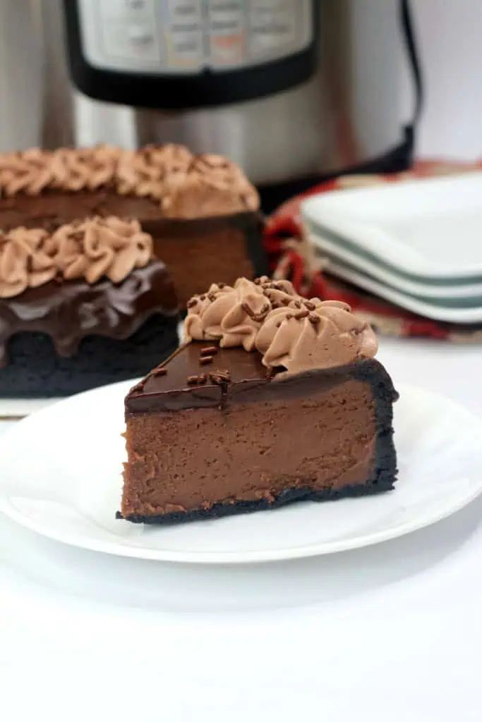 Slice of Instant Pot Nutella Cheesecake on a white serving plate with the rest of the cake in the background together with stacked plates and Instant Pot cooker.